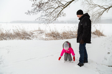 Father with daughter dressed warmly and in warm gloves builds a snowman in a snowy near the lake.
