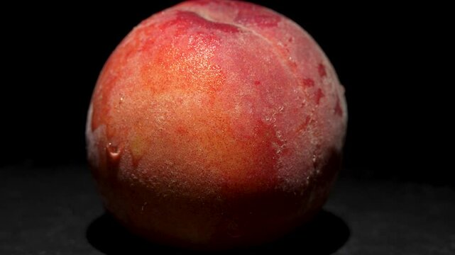 Single peach on a black background. A juicy, ripe peach rotates slowly in the light. The velvety skin has a slight sheen and displays reddish-yellow color gradients.