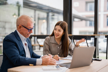 Senior businessman and young businesswoman discussing strategy during a meeting in a modern office, analyzing financial charts and using a laptop
