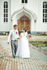 A bride and groom are walking down a brick path in front of a church. The bride is wearing a white...