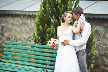 A bride and groom are standing on a green bench, with the bride holding a bouquet. Scene is...