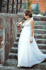 A bride is walking down a stone staircase in a white dress. She is holding a bouquet of flowers and...