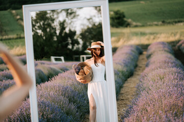 Woman in white dress poses with basket in lavender field reflected in mirror on sunny day