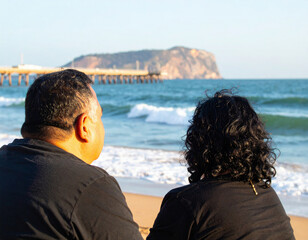 Fototapeta premium A couple sits on a sandy beach, gazing at the ocean waves and distant cliffs, enjoying a peaceful moment together.