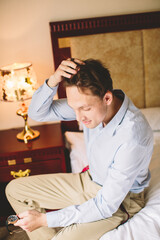 Caucasian man sitting on bed and holding wristwatch while fixing hair — modern masculinity,...