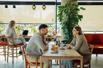 Young couple talking seriously at cafe table, woman working on laptop in background