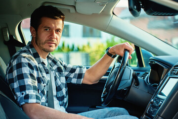 Portrait of man in shirt behind steering wheel looks at camera while sitting in modern vehicle. Person driving car