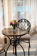 Decorative table with roses in a vase and chair in the hotel lobby. Large panoramic window with curtains and view of the pool. Summer recreation and tourism concept.