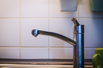 Old chrome kitchen faucet with dripping water drops captured in closeup against a white ceramic tiled wall background showing plumbing detail and water waste concept.