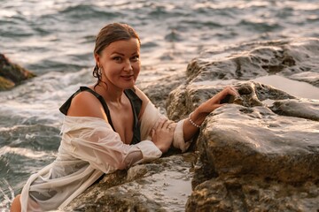Woman Beach Rocks Sunset - A woman in a white shirt and black swimsuit smiles while sitting on rocks by the ocean at sunset.