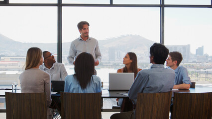 Businessman Making Presentation To Colleagues Using Laptops Sitting Around Boardroom Table