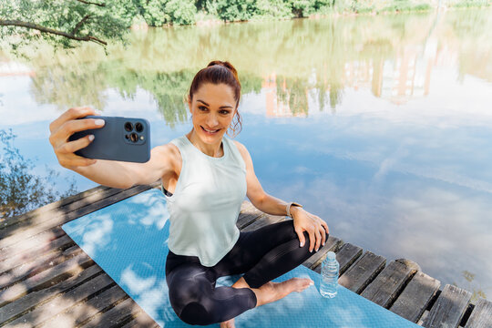 Fitness influencer capturing post-yoga selfie on wooden pier, water bottle beside, lake landscape surrounding relaxed female athlete