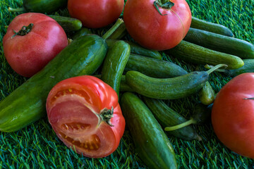 A close-up image of fresh red tomatoes and green cucumbers laid out on vibrant artificial grass. The scene evokes freshness, summer, and healthy eating.