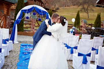 A bride and groom are kissing in front of a blue arch. The bride is wearing a white dress and the...