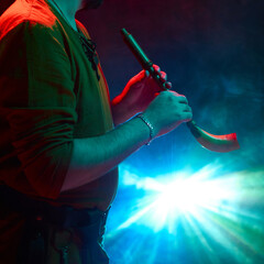 A musician plays the traditional horn under vibrant stage lighting, casting colorful patterns....