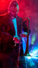 A musician performs with a trombone under vibrant stage lights and atmospheric smoke effects....