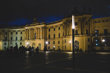 Fototapeta premium Night view of Bebelplatz square in Berlin, Germany, featuring illuminated historic buildings, silhouettes of people walking, and atmospheric city lights.
