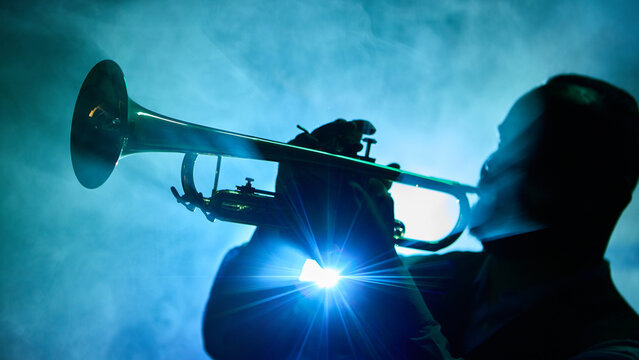 A silhouette of a musician playing a trumpet emerges against a blue smoky background. Concept of jazz or blues performance ambiance.