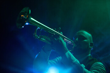 A musician plays the trumpet on a dimly lit stage with blue and green spotlights and atmospheric...