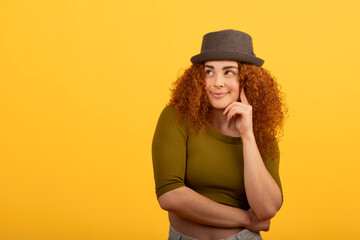 Pensive redhead woman with hat touching chin on yellow background
