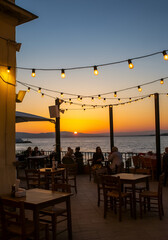 A seaside restaurant terrace with empty tables in the foreground and people sitting in the back watching the sunset under warm hanging string lights.