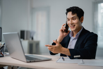 Young happy businessman wearing suit is making a phone call and gesturing while working on a laptop...