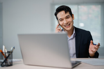 Smiling Asian businessman engaging in a phone conversation while working on a laptop in a bright office, holding a pen in his other hand, exuding professionalism and cheerfulness