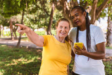 Elderly multiracial female friends smiling, sharing smartphone screen while pointing at park...
