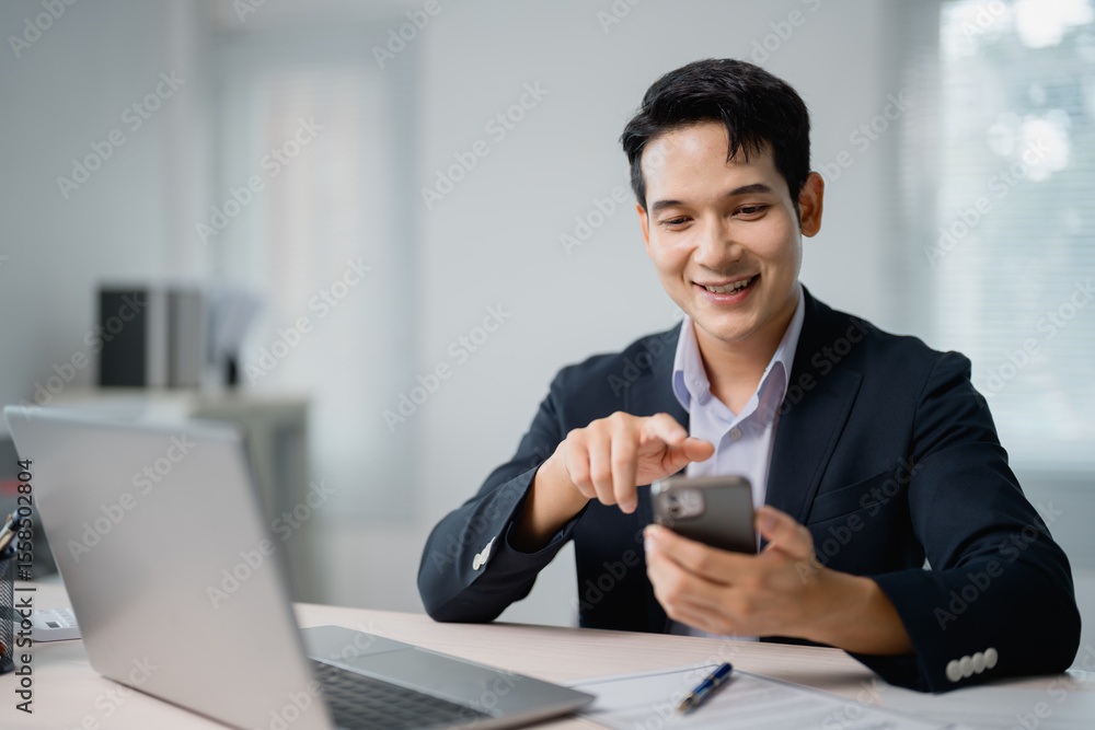 Poster smiling asian entrepreneur browsing the internet on a smartphone while sitting at a desk with a lapt