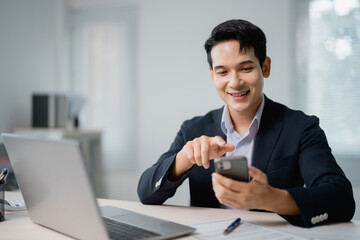 Smiling Asian entrepreneur browsing the internet on a smartphone while sitting at a desk with a...