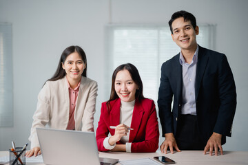 Three Asian businesspeople smiling and collaborating around a laptop in a modern office, exemplifying teamwork and unity in a bright and professional workspace