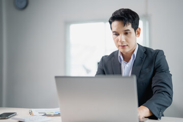 Young entrepreneur wearing suit and tie using laptop while working at desk in bright office, concentrating on computer screen with documents and smartphone nearby