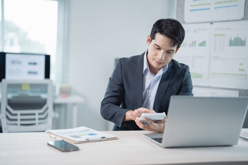 Young businessman is sitting at his desk in a modern office, using a calculator to analyze...