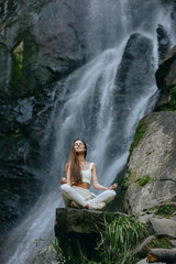 young woman meditating in lotus pose near waterfall wearing white activewear surrounded by rocks...