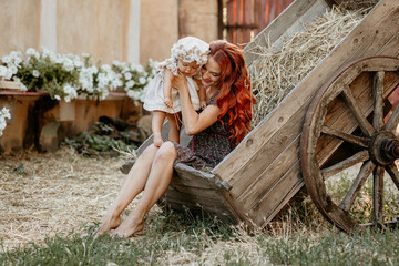 Obraz premium Happy young mother with baby in her arms sits in cart at sunset in village in summer. Girl with red hair. Motherhood. Mother's day. Mom and daughter