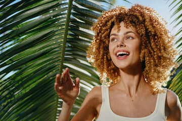 Smiling woman with curly hair enjoying sunny tropical outdoors surrounded by green palm leaves,...