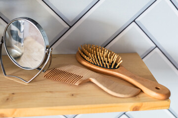 Wooden hairbrush and comb alongside round mirror on wooden shelf in modern bathroom setting