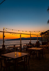 A seaside restaurant terrace with empty tables in the foreground and people sitting in the back watching the sunset under warm hanging string lights.