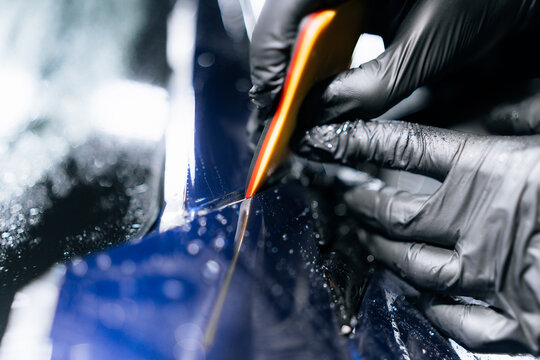 Close-up of gloved hands applying paint protection film (PPF) to a blue vehicle with precision cutting tool during a professional detailing and wrapping process in a workshop environment