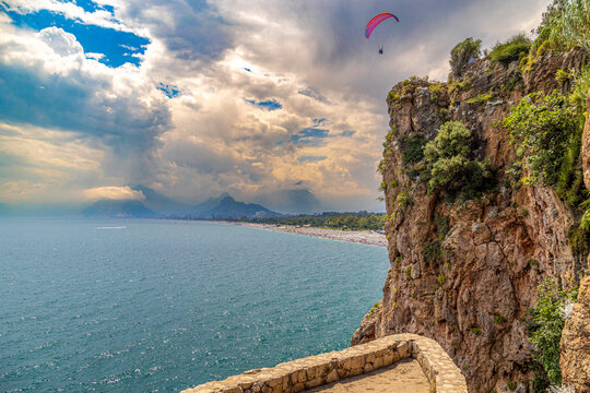 Turquoise Mediterranean Sea meets Konyaaltı Beach with paragliders above cliffs, crowded shore, and hazy Taurus Mountains backdrop. Antalya, Turkey.


