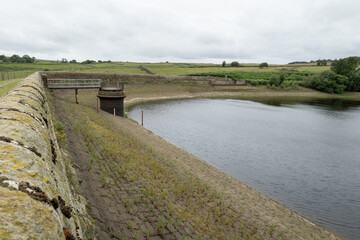 A depleted reservoir in the Yorkshire countryside shows low water levels exposing stone embankments...