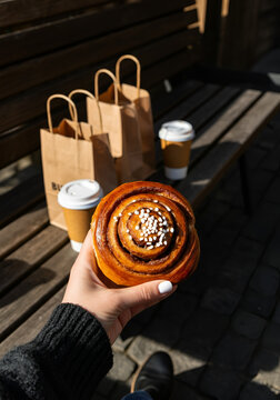 A slightly tilted top-down photo of a hand holding a Swedish cinnamon bun topped with pearl sugar. Set in a rustic outdoor caf&eacute; environment with a wooden bench, kraft paper bags, and takeaway coffee.