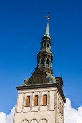 St. Nicholas Church (Niguliste kirik) tower in Tallinn Old Town, Estonia, with historic Gothic copper spire, white stone walls and medieval architecture under bright blue sky.