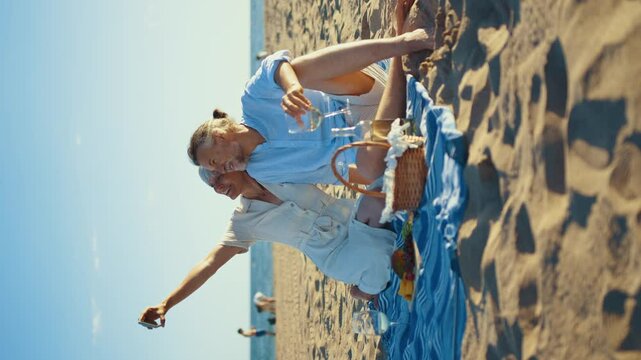 Retired couple sharing intimate moment while pouring white wine, sitting on beach blanket with picnic basket, enjoying fresh fruits during golden sunset by ocean shore