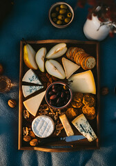 op-down photo of a rustic cheese board with pear and melon slices, assorted cheeses, olives, nuts, and crackers, arranged on a wooden tray over a textured blue picnic blanket.