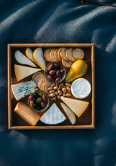 op-down photo of a rustic cheese board with pear and melon slices, assorted cheeses, olives, nuts, and crackers, arranged on a wooden tray over a textured blue picnic blanket.