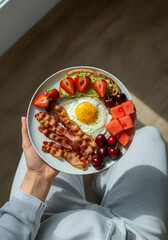 Top-down photo of a breakfast plate with crispy bacon, seasoned fried egg, buttered toast, strawberries, cherries, and watermelon cubes, held by a woman in light-colored workout clothes. 
