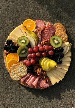 Top-down view of a vibrant charcuterie board with sliced cheeses, kiwi, grapes, orange slices, pears, blackberries, pecans, crackers, and cured meats arranged in a radial composition.