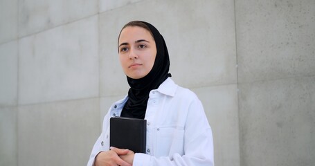 Muslim woman wearing hijab, white shirt, holding book close, gazing pensively near concrete wall,...