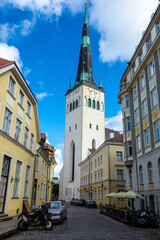 Fototapeta premium Street view of St. Olaf's Church in Tallinn, Estonia, featuring the tall spire rising above historic pastel-colored buildings on a cobblestone street.
